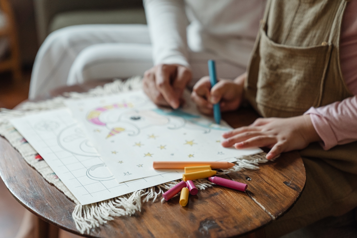 Daycare teacher sits by child coloring at a table.