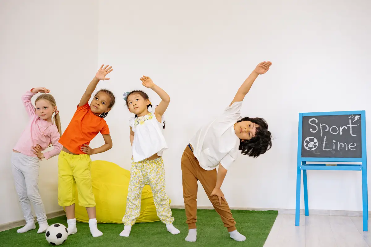 Four young children stretch their muscles in an exercise class next to a chalkboard sign that says sport time.