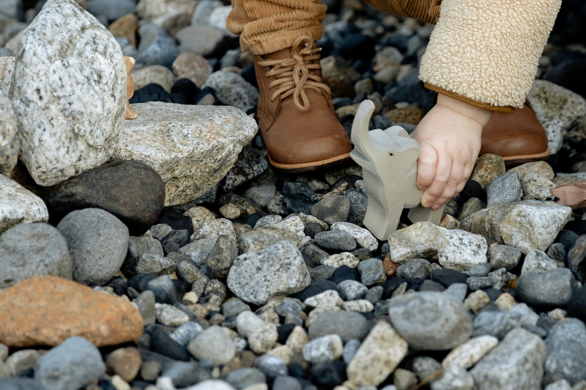 Toddler stands on a rock path and plays with a wooden toy elephant.