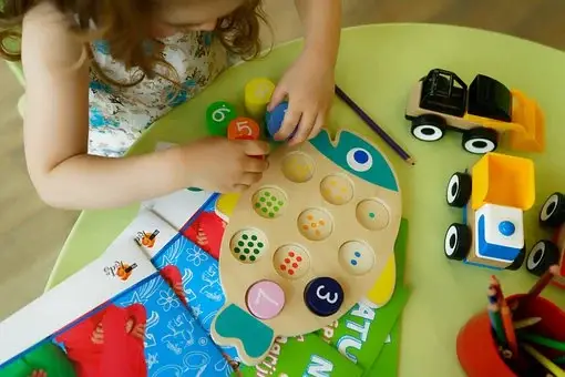Girl sits at a table playing with a wooden numbers toy with several learning activities, colored pencils, wooden toy cars.
