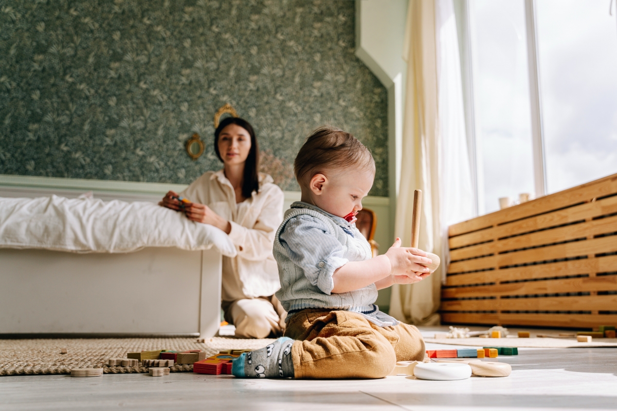 Mom sits back and watches as baby kneels on the floor and plays with a wooden stacking toy.