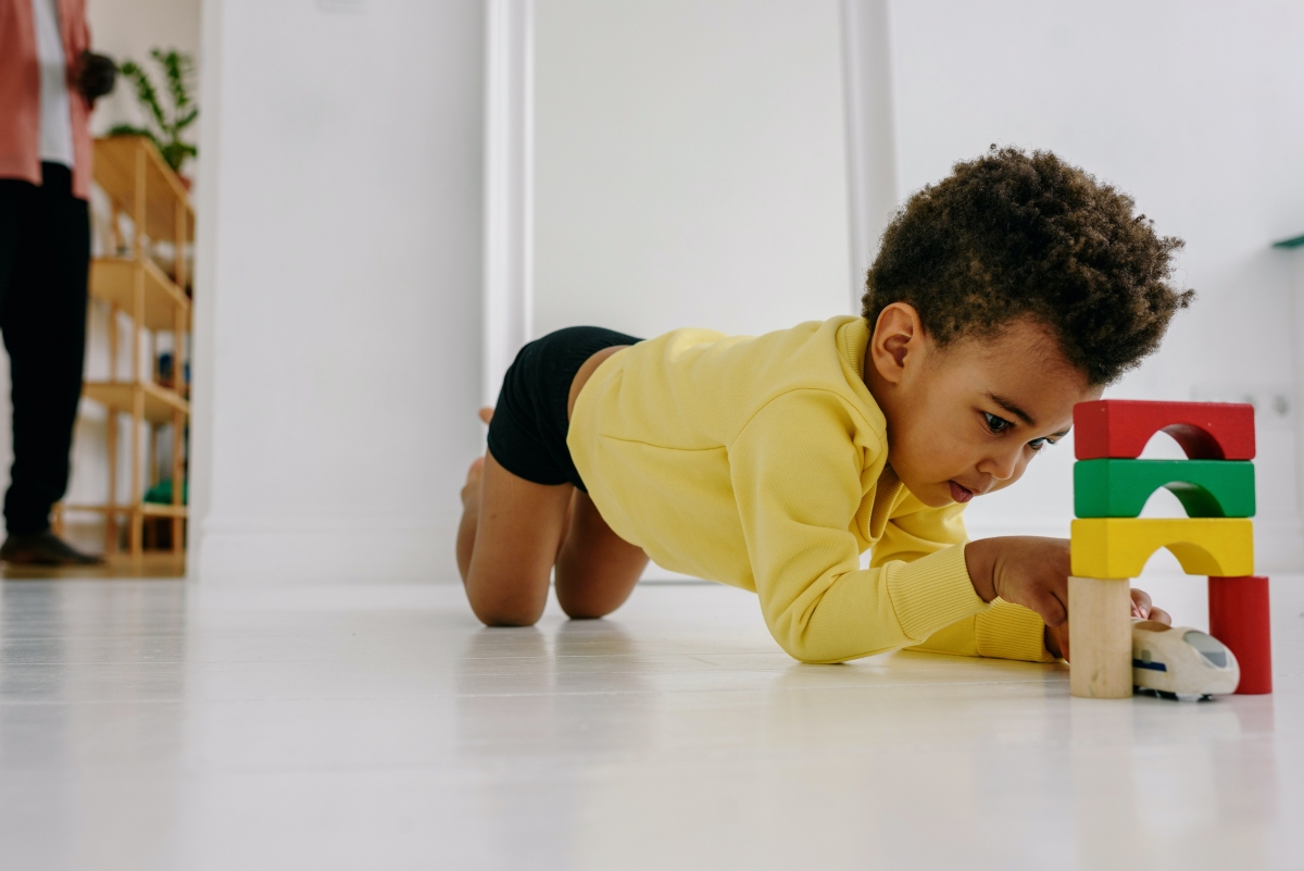 Boy kneels on the floor playing with a wooden train and wooden tower as his dad watches in the background.