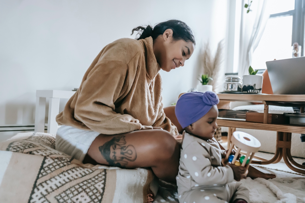 Mom smiles and watches as her baby sits and plays with a wooden toy.