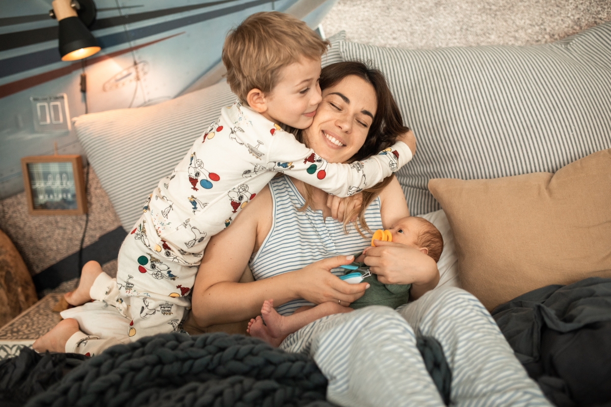 Mom smiles leaning up in bed as her young son hugs her while she holds her newborn.